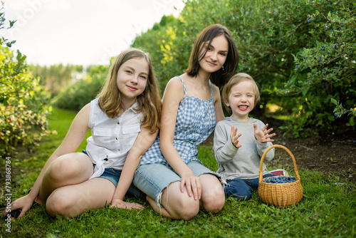 Cute teenage sisters and their little brother picking fresh berries on organic blueberry farm on warm and sunny summer day. Fresh healthy organic food for kids. © MNStudio