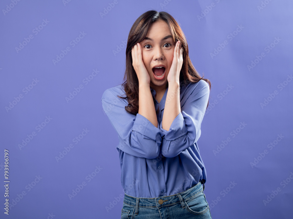 An Asian woman in a blue shirt and jeans covers her ears and screams with an angry expression against a solid blue background An Asian woman in a blue shirt and jeans covers her ears and screams with an angry expression against a solid blue background
