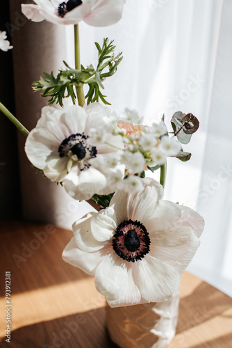 white flowers in a white vase stand on a wooden table near the window on a sunny day