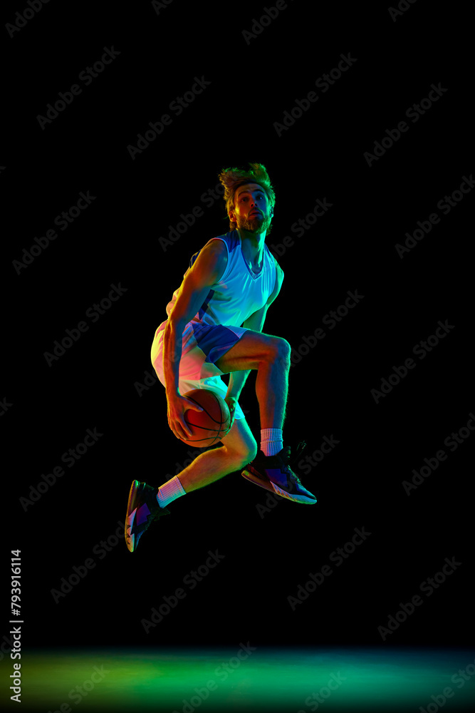Fototapeta premium Slam dunk pose. Young, focused man, basketball player in motion, training against black background in neon light. Concept of sport, competition, active and healthy lifestyle, game