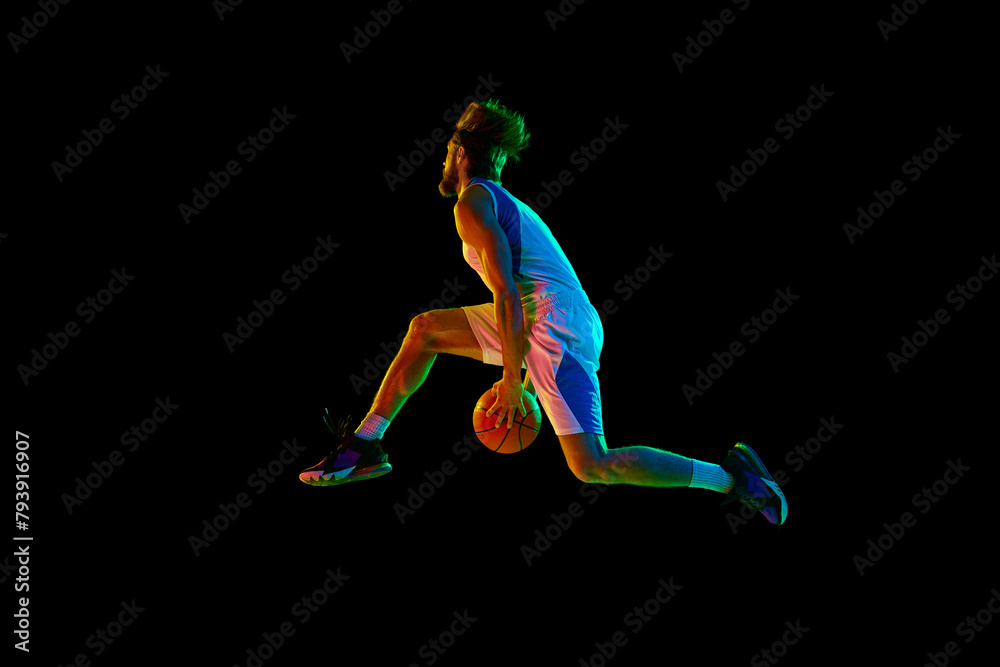 Competitive young man, basketball player in mid-air, training, jumping with ball against black background in neon light. Concept of sport, competition, active and healthy lifestyle, game