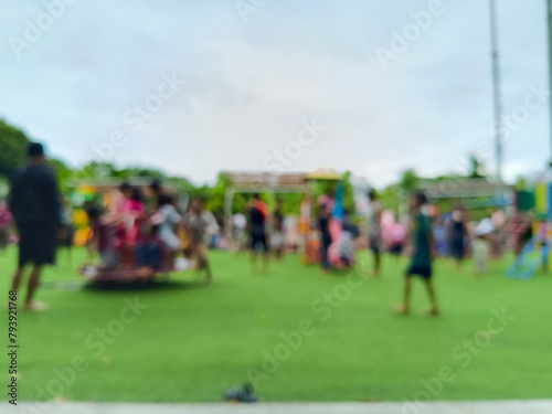 blurred, wide-angle view of an outdoor playground or park with a green grass field, featuring children and adults enjoying various recreational activities and play equipment under a bright, cloudy sky