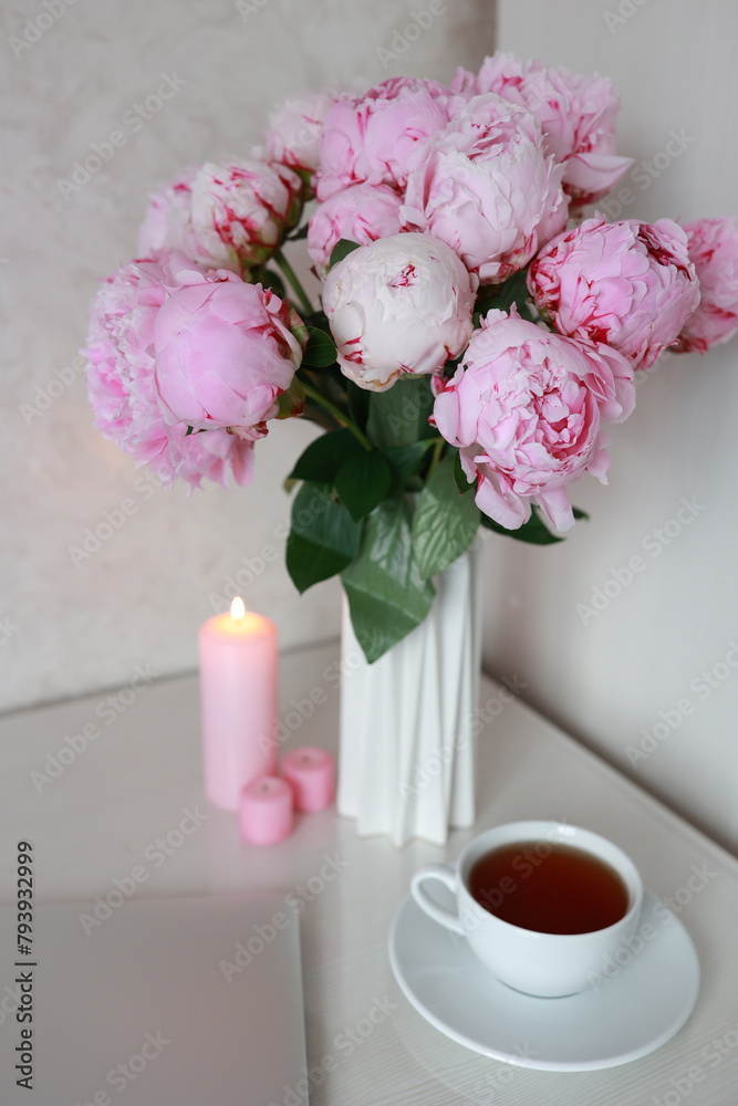 tea in a white cup, pink peonies, flowers and candles of pink color on a white background.Laptop, beautiful pink peony bouquet and notebook on white table background. minimalist home workspace. 