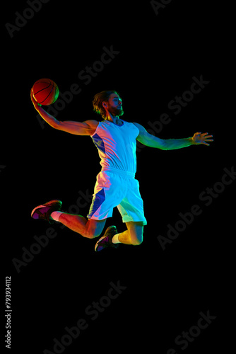 Full-length image of young men, basketball player in jump with ball, scoring goal against black background in neon light. Concept of sport, competition, active and healthy lifestyle, game