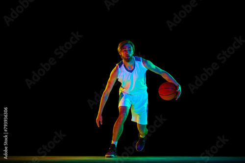 Young athletic man, basketball player in motion with ball, training against black background in neon light. Concept of sport, competition, active and healthy lifestyle, game