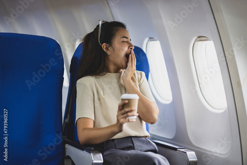 Young Asian female tourist traveling alone on a plane is drinking hot coffee and looking out the window. Woman passenger yawning and sleepy in airplane.