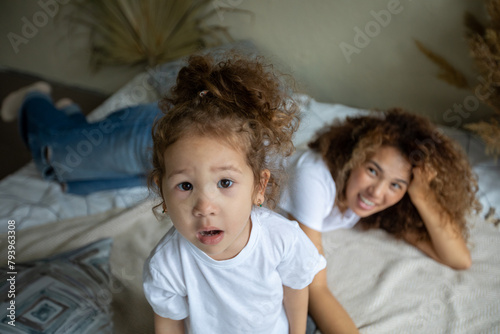 Smiling young Caucasian mom lying on comfortable bed have fun engaged in funny game activity with small daughter, overjoyed mother and little girl child playing together in cozy bedroom at home