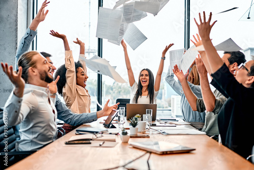 Celebrating success. Joyful command throwing up documents in air above long wooden table with excited expression. Playful business people reaching goal and entertaining together as sign of victory. photo