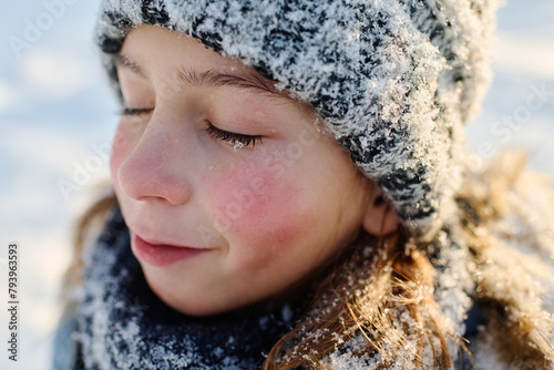 Close-up portrait of cute boy with closed eyes and snowflake on eyelashes standing in front of camera on winter day and enjoying stroll