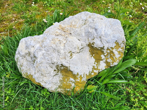 Großer, unregelmäßig geformter Stein, der auf einem Grasfeld liegt. Der Stein hat eine raue, unebene Oberfläche mit Flecken von Moos, die ihm eine kontrastreiche graue Farbe verleihen.