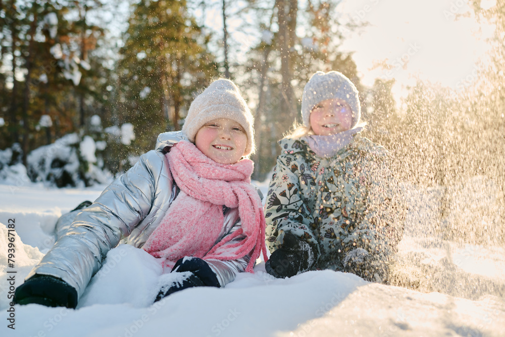 Two cheerful girls in winterwear lying in snowdrift and looking at camera while throwing snow and enjoying sunny winter day in park