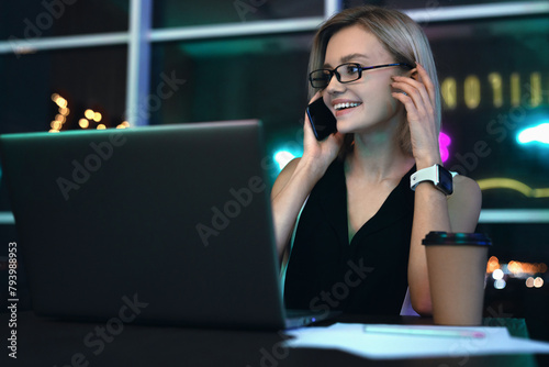 Cheerful businesswoman talking on cellphone sitting at table and working. Caucasian female sitting at office late night and having telephonic conversation with client and smiling with look away photo