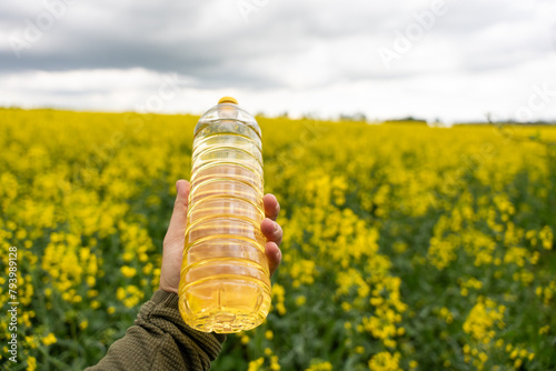 A bottle of rapeseed oil in a hand against the background of a yellow blooming rapeseed field. A bottle of rapeseed oil and blooming rapeseed fields.