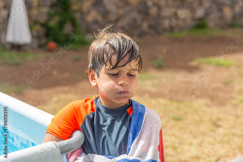 Contemplative young boy wrapped in a towel by the swimming pool, wet hair, summer day. photo