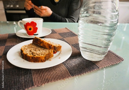 Frau sitzt vor Küchentisch mit Karaffe Wasser, Tasse Kaffee und Bananenbrotscheiben auf weißem Teller in Küche 