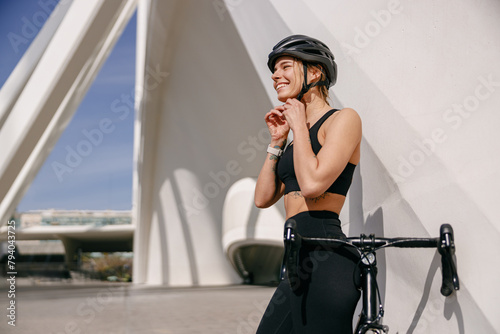 Smiling female cyclist is putting helmet on head before going to go ride by bicycle in city