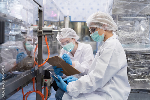 Two female worker wearing uniform, hairnet and protective mask working inside automated conveyor belt production line in industry factory