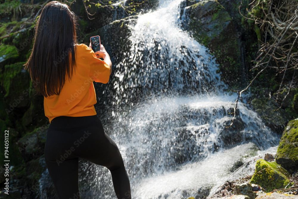 Fototapeta premium Woman standing in front of waterfall and taking pictures with the phone 