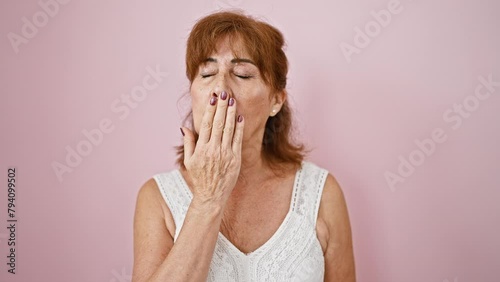 Exhausted middle-aged woman languidly yawning; laziness captured over isolated pink background. boredom visible in every aspect of tired expression and drooping hand covering mouth. photo