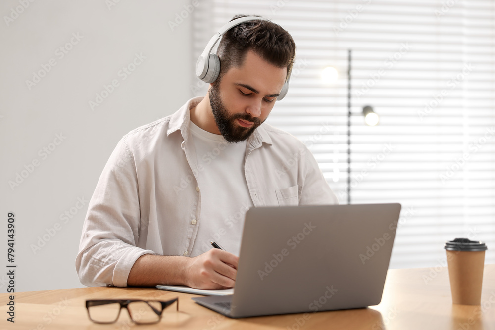 Young man in headphones writing down notes during webinar at table in room Young man in headphones writing down notes during webinar at table in room