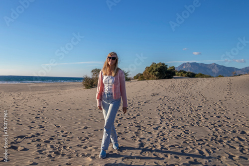 Happy vivacious young woman giggling and smiling as she tries to stop her long blonde hair blowing about her face on a sandy beach in autumn