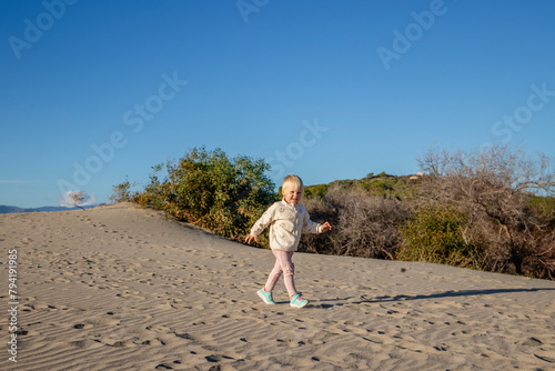 A happy 2-year-old girl runs along the sand dunes next to the sea.
