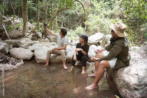 Group of three Asian friends, two men and one woman, have a good relationship, hiking and resting at the stream, nature activities, camping in the forest, summer time trip, vacation Lifestyle concept
