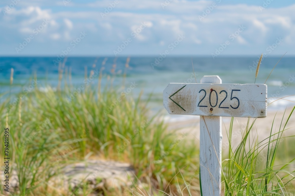 A white sign with the number 2025 on it is standing in a field next to the ocean