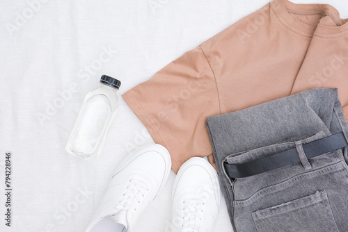Unisex clothes and accessories set on white background top view: black jeans, brown tee shirt, white trainers, bottle of water. modern and casual outfit. fashion, shopping, leisure. 