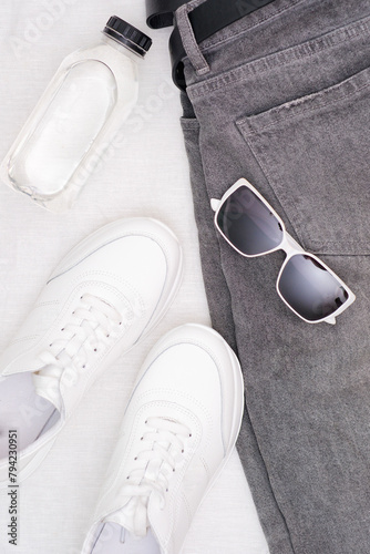 Unisex clothes and accessories set on white background top view: black jeans, sunglasses, white trainers, bottle of water. modern and casual outfit. fashion, shopping, leisure. 