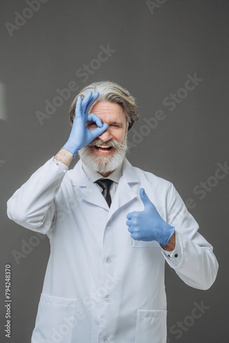 Portrait of an expressive cheerful senior doctor in a white coat on a gray background.