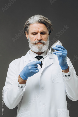 Gray-haired senior doctor in white coat holding syringe with injection, isolated on gray background.