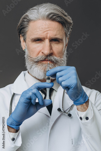 Portrait of a senior doctor in gloves and medical gown holding a syringe. isolated on gray background.