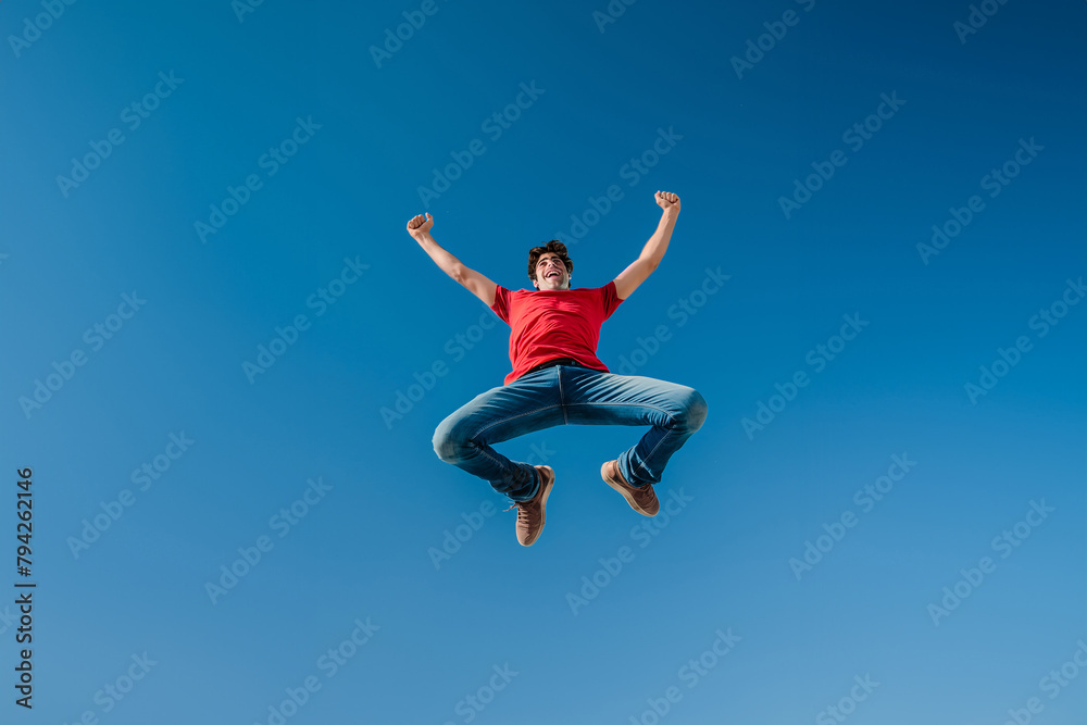 Joyful man in red shirt celebrating with a high jump against clear blue sky
