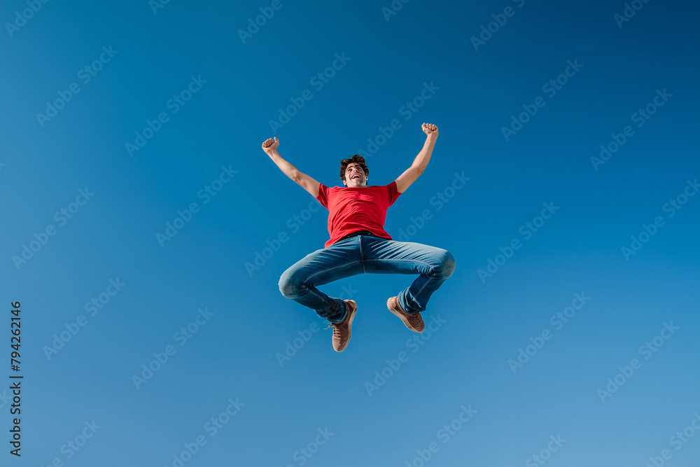 Joyful man in red shirt celebrating with a high jump against clear blue sky Joyful man in red shirt celebrating with a high jump against clear blue sky