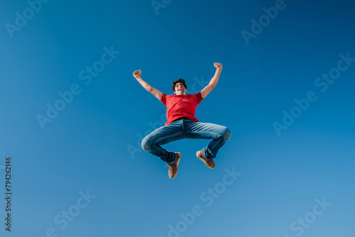 Joyful man in red shirt celebrating with a high jump against clear blue sky