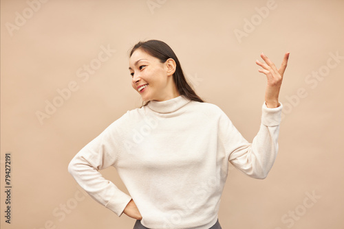 Charming charismatic asian female in beige mockup turtleneck raising her hand up and gesticulating like acting or rehearsing her part, holding right hand on waist looking aside against pink background