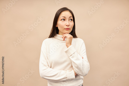 Portrait of beautiful asian woman standing against pink background in beige turtleneck, touching her chin and looking upwards as if hatching an idea or contemplating decision or plan