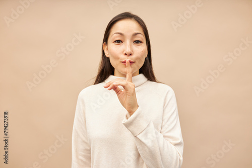Studio portrait of cute asian woman in turtleneck showing shh gesture pressing index finger against lips, sharing secret recommendations of special offer of discounts, standing against pink background