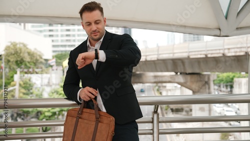 Happy business man looking watch while waiting his colleague for working together at bridge. Caucasian manager looking at time while standing in suit surrounded by urban city view. City life. Urbane.