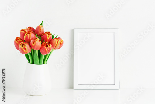 White frame mockup and red tulips in a white vase on a white table by the wall