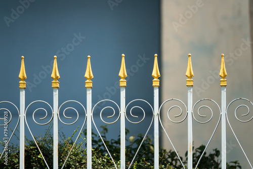A fence with gold spikes on it. The fence is white and has a blue background