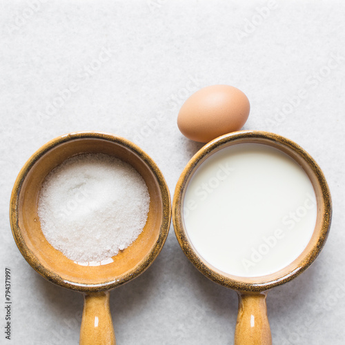 Overhead view of mise en place of ingredients for making custard, egg sugar and milk in ceramic ramekins, process of making custard