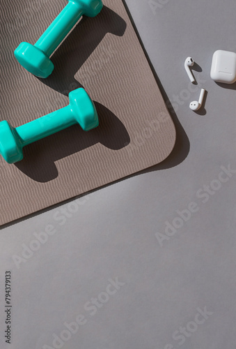 Sport or workout concept flat lay with blue dumbbells, yoga mat and white wireless headphones on the grey background. Copy space