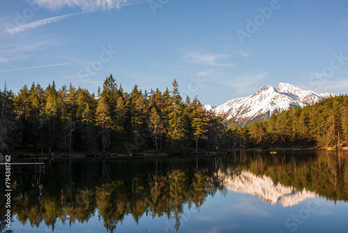 Snow-capped mountain reflected in the still waters of a forest-fringed lake under a clear blue sky.