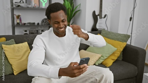 An exuberant young man celebrates with a fist pump while using a smartphone in a modern living room. photo