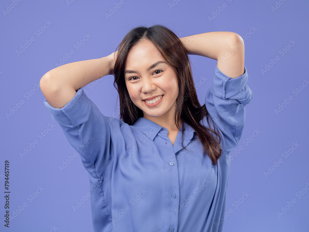 Portrait of an Indonesian Asian woman, wearing a blue shirt, smiling with her hands behind her head, isolated against a purple background Portrait of an Indonesian Asian woman, wearing a blue shirt, smiling with her hands behind her head, isolated against a purple background