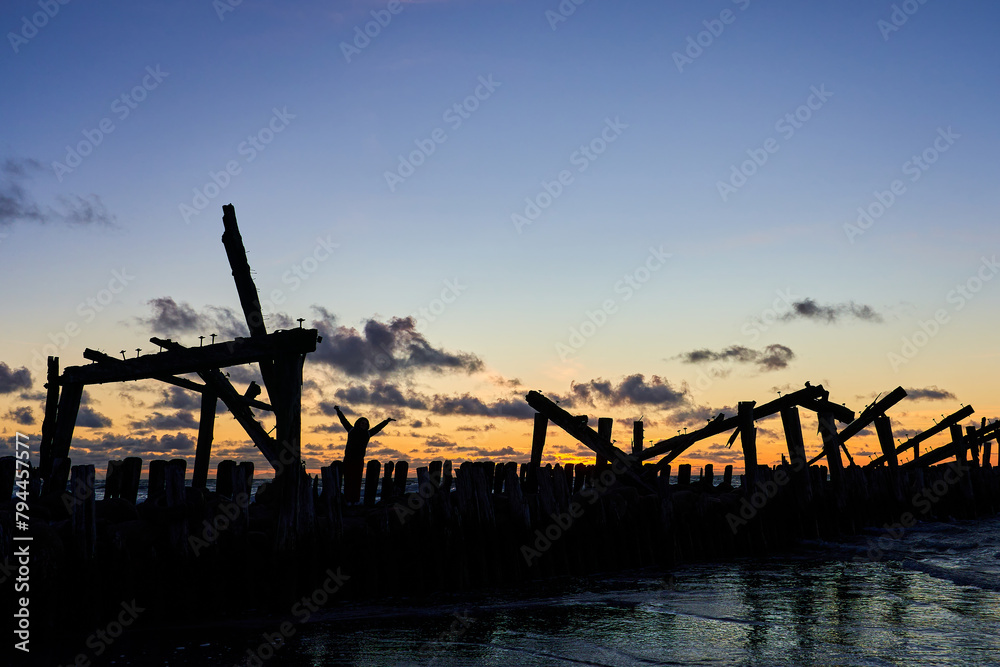 The silhouette of a man among the ruined old pier in the background light against the sunset The silhouette of a man among the ruined old pier in the background light against the sunset