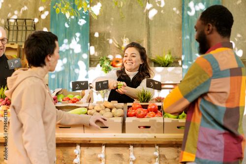Boyfriend and girlfriend tasting fresh green apple before purchasing organic fruits and vegetables at farmers market. Happy farm produce stand owner offering free samples, bio natural products.