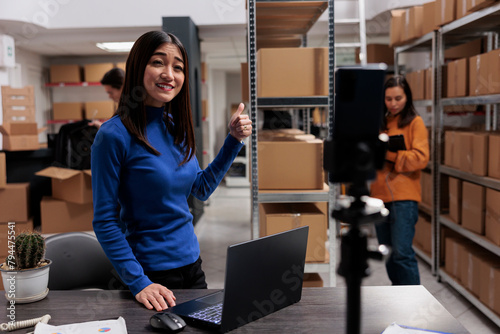 Logistics and distribution manager advertising merchnadise in storehouse on smartphone camera. Warehouse supervisor streaming employees team working in storage room together photo